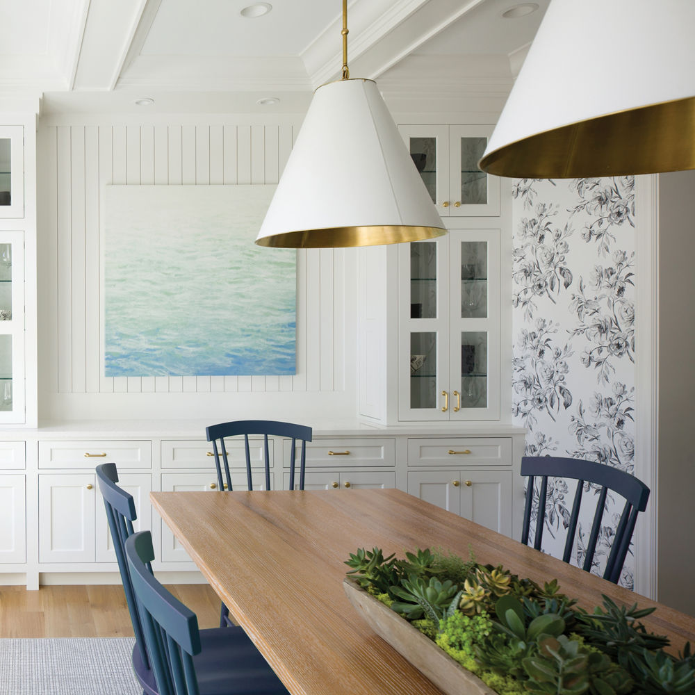 A coastal inspired dining room with blue chairs, white hutch topped with white quartz, a sea-inspired painting, and wooden flooring.